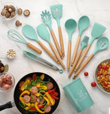 Silicone kitchenware utensils set in teal color with wooden handles, arranged with cooked food and ingredients on a marble countertop.