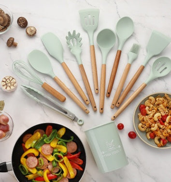 Set of mint green silicone kitchen utensils with wooden handles, featuring spatulas, whisk, and tongs, on a countertop with food.